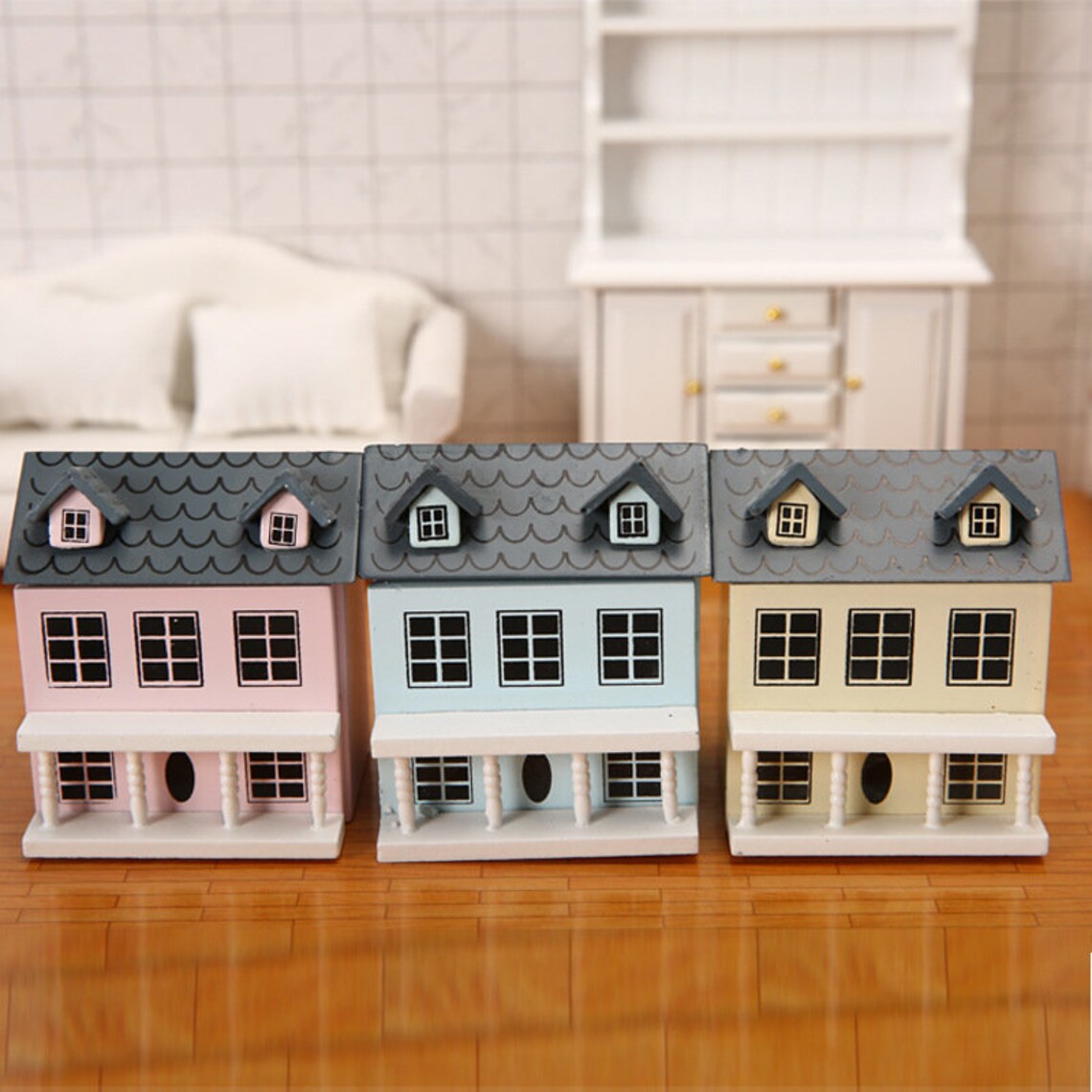 A single lycheepuzzles 1:12 Dollhouse Miniature Wooden Little House with a blue roof is showcased on a wooden surface, emphasizing its detailed construction. In the background, there's a white sofa and cabinet, enhancing the overall appeal.